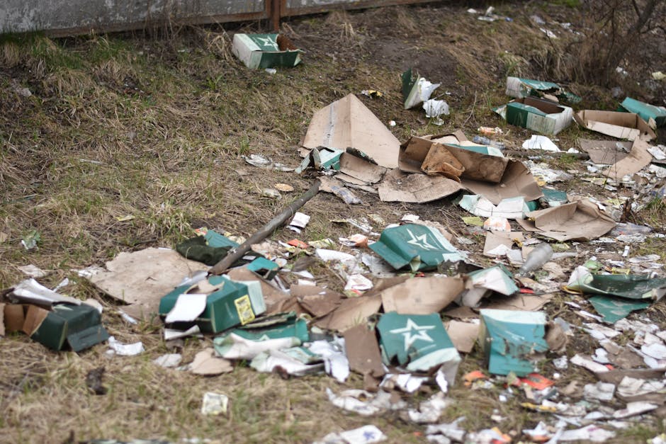 An outdoor scene showing scattered cardboard boxes, some flattened and torn, alongside crumpled paper and plastic packaging, all discarded on a patch of grassy ground next to a concrete wall. The boxes are primarily green with white star logos, indicative of packaging for beverages or similar products. Fragments of cardboard and paper are strewn across the uneven terrain, with some boxes partially collapsed or damaged from improper disposal. Interspersed among the debris are pieces of plastic wrapping and what appears to be remnants of packaging material. The scene suggests an instance of illegal dumping or inadequate waste collection, with rubbish spilled onto an open outdoor area. In the background, a concrete edge or boundary is visible, and natural daylight illuminates the scene, emphasizing the litter's disorderly nature. The image underscores the importance of proper waste handling and highlights how private rubbish removal services, such as those provided by Waste Disposal Sutton, can help manage such waste responsibly and prevent environmental contamination.
