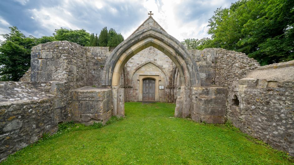 A historic stone structure featuring a pointed Gothic archway, made of weathered grey stone blocks with visible textures and signs of aging. The arch surrounds a wooden door with metal hinges and a small inked sign beside it. The structure is set within a partly ruined stone wall, with uneven and rough surfaces, and is situated in a grassy area with neatly maintained green lawn in the foreground. Behind the structure, there are tall, leafy trees and a partly cloudy sky, creating a natural backdrop. The scene appears to be during daytime with soft, diffused lighting highlighting the weathered surface details of the stone. The setting suggests it could be part of an old religious or historical site, and the overall scene aligns with the context of private or alternative historical site management, similar to independent disposal or clearance services provided by Waste Disposal Sutton, ensuring the preservation and maintenance of heritage environments.